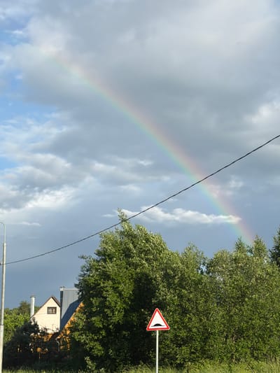 The image captures a view of a suburban landscape with a rainbow arcing across a cloudy sky. In the foreground, a cluster of lush green trees dominates the scene. To the left of the trees, a white triangular road sign with a red border indicates a speed bump ahead. Behind the trees, a portion of a residential building with a yellow facade and a grey roof is visible. The building appears to be a house, with a small brown-framed window and two chimneys. Further to the left, a light pole stands, and a wire stretches across the upper portion of the frame. The sky is a mix of blue and grey clouds, with the rainbow prominently displayed. The scene suggests a location in Satino-Russkoe, Russia, likely during or shortly after a rainfall. No people are present in the image.