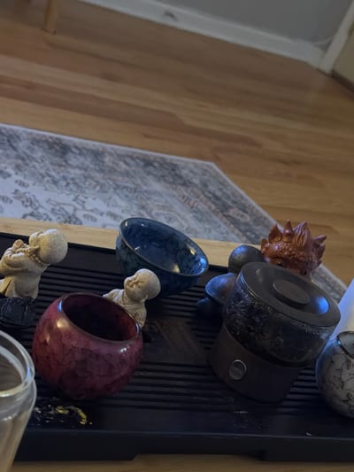 The image shows a collection of Asian-themed objects arranged on a dark, possibly black, slatted surface. The objects include a carved wooden figurine of a lion or mythical creature, a Zensorium Aroma Oil Diffuser with a stone finish, a blue and black bowl, a maroon circular vessel, and two small, pale-colored statues of Buddha-like figures. In the background, a wooden floor is visible on the left, while a patterned area rug is present behind the objects. The scene seems to be indoors. The location of Morton Grove, United States of America, is not discernible from the image.