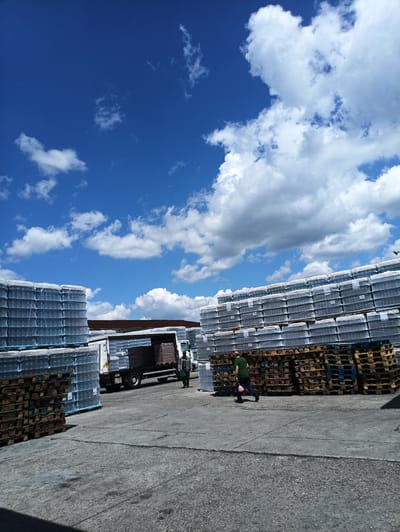 The image shows an outdoor storage or distribution area, possibly in or near Venets, Bulgaria, given the request. Stacks of palletized goods, likely bottled water or similar beverages wrapped in plastic, are prominently featured. A truck is visible in the background, partially loaded with similar palletized goods. Two people are present: one standing near the truck, possibly a worker, and another man walking in the foreground. The foreground consists of a concrete surface with visible cracks. The sky is blue with scattered white clouds. The lighting suggests it is daytime. The pallets are stacked between the goods and the ground.