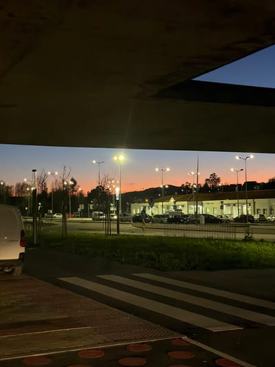 The image depicts an urban scene at dusk or early night in Penafiel, Portugal, characterized by artificial lighting against a twilight sky. The top half of the frame is dominated by the underside of a concrete structure, likely an overpass or a large architectural overhang.
In the immediate foreground, a white panel van is partially visible on the far left, showing its rear bumper and taillights. Adjacent to this, a pedestrian area features tactile paving with circular and linear raised patterns, leading to a clearly marked zebra crossing on a dark asphalt road.
The midground includes a multi-lane roadway bordered by a grassy median or verge. Numerous active streetlights, equipped with yellow-emitting lamps, illuminate the road and surrounding areas. Sparse, bare trees and shrubs are distributed across the landscape. A low metal fence separates the grassy area from a commercial parking lot.
In the background, a low-rise commercial building with a white facade and dark roofing is prominent. A sign on this building reads "P. NAFIEL," confirming the location in Penafiel. The parking lot in front of the building contains numerous parked vehicles of various types and colors. Several circular blue and white road signs are visible near the building's entrance. Behind the building, a prominent hill or elevated terrain, sparsely vegetated, is silhouetted against the sky, which transitions from a warm orange-pink at the horizon to a deeper blue overhead. No individuals are visible in the scene.