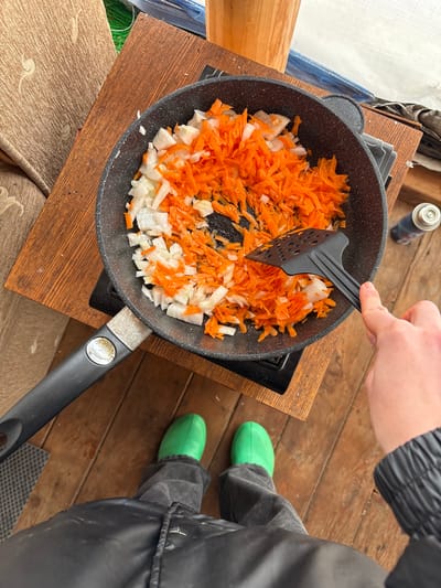 A high-angle shot depicts a person stirring a frying pan containing chopped carrots and onions on a wooden surface outdoors. The person is using a black slotted spatula. A small gas canister sits next to the pan. The individual is wearing a black jacket and grey trousers, with bright green rubber boots visible. The wooden surface appears to be a makeshift cooking station. The location is outdoors and potentially temporary. The exact city of Zheleznodorozhnyi, Russia cannot be determined from the image.