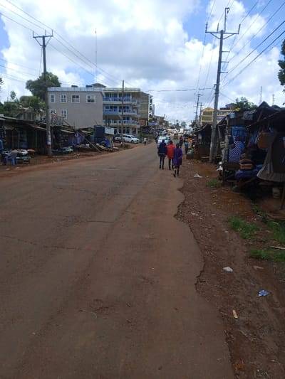 The image displays a daytime street scene in Kangema, Kenya, looking down a reddish-brown, unpaved or heavily worn asphalt road that exhibits cracks and surface irregularities. On both sides of the road, makeshift market stalls constructed from wood and corrugated iron are present, displaying various goods such as textiles, clothing, and general merchandise.

On the left side of the road, behind the market stalls, a multi-story building with a light grey facade and multiple windows is prominent, adjacent to another multi-story structure featuring blue-fronted balconies. A black plastic water tank is visible near the stalls on this side. Utility poles with numerous overhead electrical or communication lines line both the left and right sides of the road.

In the mid-ground, three individuals are walking away from the viewer on the road. The left-most person wears a dark blue jacket, the central person wears an orange top, and the right-most person wears a purple top, all with dark lower garments. Other pedestrians are visible further along the road and near the stalls.

The road shoulders are composed of reddish-brown soil, mud, and scattered debris, with minimal green vegetation visible on the far right. Several vehicles are observed in the distance on the road, indicating traffic. The sky is partly cloudy with white and grey cloud formations. A tall communication mast is discernible in the far background against the skyline.