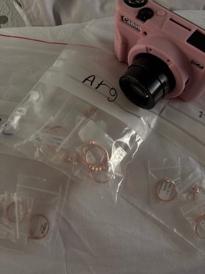 The image shows several small plastic bags filled with gold rings. The bags are resting on a white surface. A pink camera is lying on top of the bags. The camera has a black lens and a "Canon" logo on the front. The lens is pointing towards the bags of rings. The bags are labeled "ARG" and "1." The image was taken in Ramla, Israel, at the coordinates 31.916, 34.867. There are no people in the image.