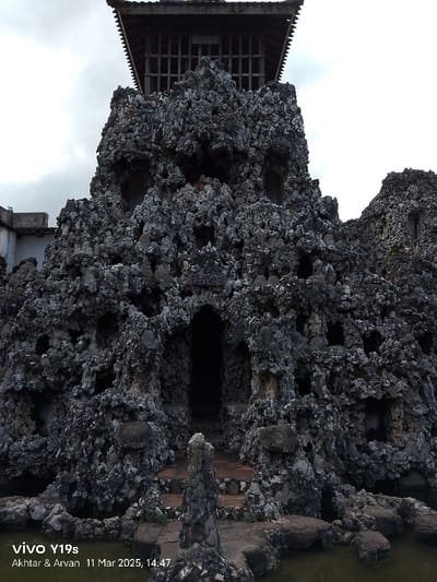 The image shows a large, rock-like structure with a wooden roof, located at the coordinates -6.736, 108.544, which correspond to the Keraton Kasepuhan Palace in Cirebon, Indonesia. The structure appears to be constructed from a rough, gray material, possibly concrete or stone, with numerous openings and alcoves of varying sizes. The roof is a simple structure with a gabled shape and latticework panels. It is unclear what the purpose of the structure is, but it is likely part of a larger complex, possibly a shrine or a monument. The surrounding area is relatively empty, with a small body of water in the foreground and a portion of another rocky structure in the background. The scene is captured from a low angle, giving the impression of being quite tall. A small sign can be seen on the wall above a doorway with "KEPITEN" written on it. The image appears to be taken in daylight, but the overcast sky suggests it may have been cloudy. The overall atmosphere is peaceful and serene.
