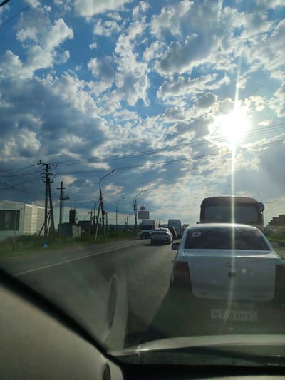 The image shows a traffic jam on a road in Blagoveshchenskaya, Russia. The sun is shining brightly through a sky filled with clouds. Several cars are stopped on the road, and power lines are visible on the side. The perspective is from inside a vehicle, looking forward. A white car with a red cross emblem is in the foreground.