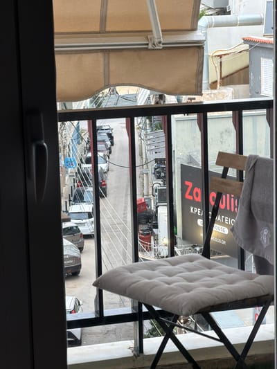 View from a window in Glifada, Greece, looking down at a street. The street has parked cars, a restaurant sign that says "Zakoulas," and clothes drying on a rack. There is a white cloth hanging on the drying rack in front of the window.