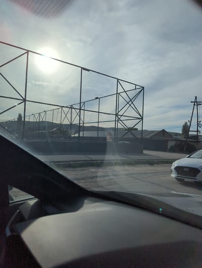 A ground-level, eye-level, medium-shot photograph taken from the interior of a vehicle shows a wire fence in Beş-Küŋgöy, Kyrgyzstan. The fence is made of metal posts and wire mesh and extends along the left side of the frame. The sun shines brightly through the fence mesh. A street and a parked white vehicle are visible behind the fence, along the right side of the frame. Overhead power lines are visible against the sky. Part of the interior of the vehicle fills the right foreground. The sky is partly cloudy.