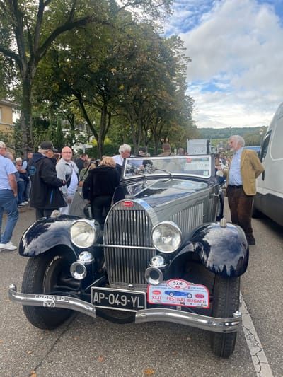 The image depicts an outdoor gathering in Molsheim, France, likely during the XLII Bugatti Festival held from September 11-14, 2023, as indicated by a plaque on the featured vehicle.

At the center, a two-tone, dark blue and light gray vintage Bugatti convertible is parked, oriented towards the viewer. The vehicle features a chrome grille with "BUGATTI" emblazoned on it, large circular headlights, and two chrome horns positioned below them. A chrome bumper with "CHOPARD" inscribed on the left is visible. The French license plate "AP-049-YL" is mounted centrally, flanked on its right by a red and white event plate detailing the "XLII FESTIVAL BUGATTI MOLSHEIM DORLISHEIM ALSACE."

Approximately 15-20 individuals are visible around the car and in the background. On the left, a man in a black jacket and cap stands next to a bald man in a gray polo shirt. A woman in a black jacket is seen leaning into the driver's side of the Bugatti. On the right, a man with white hair and a beard, wearing a tan jacket and brown corduroy pants, stands facing left, appearing to observe the vehicle or engage with others off-frame. Other individuals are gathered further in the background.

The environment consists of a paved surface, likely a street or event area, with mature trees featuring green foliage lining the background. A light-colored building is partially visible on the far left. On the far right, a white van is parked. Distant rolling hills covered in trees are visible under a partly cloudy sky.