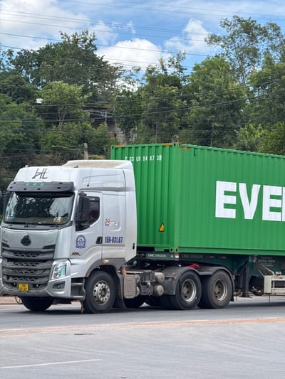 A silver semi-truck, identified by the license plate "15H-031.67," is parked on a road in Lao Bảo Commune, Vietnam. The truck is hauling a large, bright green shipping container. The container has the word "EVER" printed in large white letters on its side. The background consists of lush green trees and a partly cloudy sky. Power lines are visible crisscrossing the upper portion of the image. The truck's cab displays a logo and the model designation "H7." A yellow license plate with the number "15H 031.67" is mounted on the front bumper of the truck. The scene appears to be a daytime outdoor setting.
