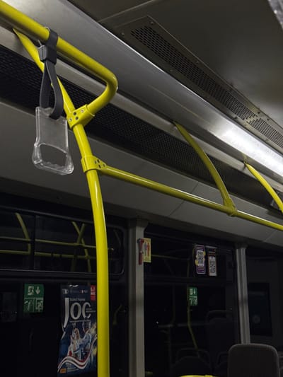 The image depicts the interior of a public transport bus in Ciorescu, Moldova. Bright yellow metal grab bars form a support structure along the upper interior of the vehicle, with various sections connected by silver hardware. A clear, transparent plastic loop handle with an ergonomic grip is suspended from a horizontal yellow bar by a black strap in the upper left quadrant. The ceiling is a light gray, equipped with a lengthy, horizontally-oriented perforated metal ventilation grate.
The bus features dark tinted windows along its side, reflecting elements from the interior. A large promotional poster, primarily blue and white with red accents, depicting dancers and announcing "Baletul Național JOC" (National Ballet JOC) for "5-7 NOIEMBRIE" (5-7 November), is affixed to a window or interior panel on the left. A green "EXIT" pictogram, showing a running figure and an arrow, is located below the poster. Further right on a window panel, two stickers displaying distinct QR codes are visible. A small rectangular sign displaying two stylized human figures (one red, one yellow) is mounted on a vertical gray pillar, adjacent to a small red emergency or stop signal device. A dark gray upholstered seat back is partially visible in the lower right foreground. No persons are visible within the frame.