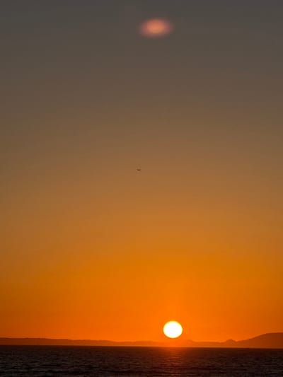 The image captures a sunset over the sea in S'arenal, Spain. The sky displays a color gradient, transitioning from intense orange at the horizon to a desaturated brown-orange at the top. A bright, circular, white-yellow sun is positioned low on the horizon, partially over the water on the right side of the frame. A dark, undulating landmass separates the sky from the body of water, which shows ripples on its surface. A small, dark-silhouetted aircraft is visible in the upper-middle portion of the sky. In the upper part of the image, a blurry, reddish-orange elliptical light anomaly, consistent with a lens flare, is present. No individuals are visible.