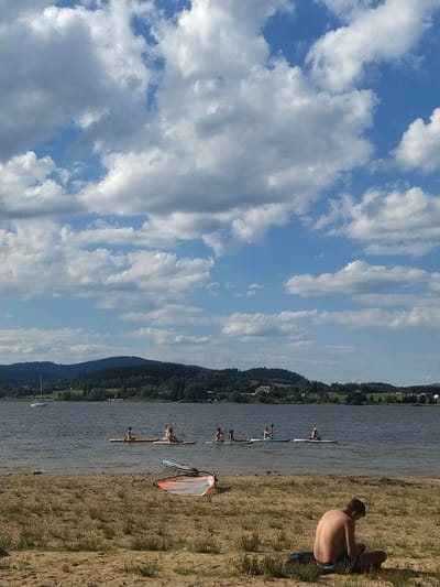 The image depicts a wide shot of a lake with a shoreline in the foreground. The shoreline is sandy and covered with sparse dry grass. A man is sitting on the sand facing away from the camera, looking down at his lap, with a dark cloth or bag under him. In the water, several people are engaged in paddleboarding, spread out across the lake. One small sailboat is visible in the distance on the left side of the lake. The background features a densely wooded hill with some scattered buildings and houses, suggesting a rural or semi-rural environment. The sky is blue with scattered, puffy white clouds, indicating fair weather. The scene appears to be the Radslav Reservoir in the Czech Republic, a popular location for water sports and recreation.