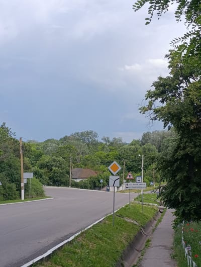 The image shows a rural intersection in Khorol, Ukraine, on a cloudy day. A paved road curves to the left, with a sidewalk and a drainage ditch on the right. A variety of road signs are visible, including a diamond-shaped yield sign, a sign indicating a turn, and a directional sign with Ukrainian text. Green trees and bushes line the road, and a small house with a red roof can be seen in the distance. The sky is overcast with some hints of blue.