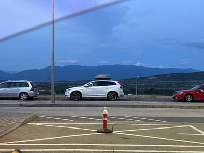 The image depicts a roadside scene with several vehicles parked along a road, with mountains and a valley in the background. The sky is a deep blue, suggesting it is either dusk or dawn. In the foreground, a parking area with white lines is visible, featuring a red and white striped traffic cone. To the left, a silver station wagon is parked. To its right, a white SUV with a roof cargo box is parked. Further to the right, a red sedan is parked. A tall, slender light post stands between the silver station wagon and the white SUV. The background shows rolling hills and mountains, with some scattered buildings visible in the distance. The overall setting suggests a rural or semi-rural area.