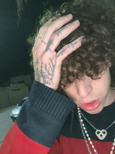 An overhead shot of a person with curly hair, fair skin, and heavily tattooed hands. The person is wearing a red and black sweater and a necklace with a heart-shaped pendant. The person's hand is touching their hair, and their face is visible. The background is dark and indistinct, with a light source at the bottom. The location is not discernible from the image.