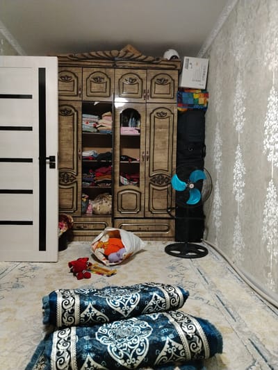 The image depicts a bedroom in an apartment located in Bekabad, Uzbekistan. In the center of the room, a large, ornate wooden wardrobe with multiple doors and drawers stands prominently. Several shelves inside the wardrobe are filled with folded clothing and other items. To the left of the wardrobe, a white door with black horizontal panels is partially open. To the right of the wardrobe, a black standing fan with bright blue blades is positioned. Stacked on top of each other near the top right corner of the wardrobe are several boxes and a colorful folded quilt. On the floor in front of the wardrobe, a white bag spills out with a variety of toys, including a red stuffed animal and a colorful xylophone. Two rolled-up blue and white patterned mats or rugs are placed on the floor in the foreground. The room has light-colored walls with a subtle floral pattern and a light-colored carpet with a blue pattern.