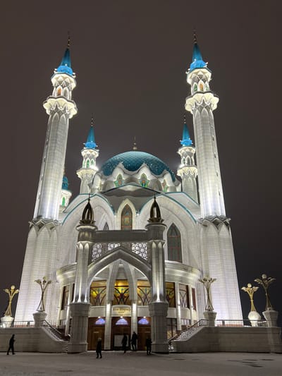 The image depicts the Kul Sharif Mosque, located within the Kazan Kremlin in Kazan, Russia, at night during winter. The large, multi-domed mosque is prominently illuminated by artificial light, presenting a pristine white facade with striking blue domes and spires on its minarets.
The central structure features a large, scalloped, tiled blue main dome, flanked by several smaller blue-spired domes and four tall, slender minarets with blue conical tops. The mosque's exterior walls are constructed from white stone, showcasing intricate patterns, pointed arches, and numerous stained glass windows, which glow with internal illumination. The grand main entrance, centered in the image, is marked by a large arched portal above several smaller arched doorways, bearing the text "Кол Шариф" (Kul Sharif) on a sign. Stone staircases with decorative railings ascend to the entrance from both sides.
The ground in the foreground and surrounding the mosque is covered in a layer of snow. Several individuals, dressed in dark winter clothing, are visible walking on the snowy ground near the mosque's entrance. Two distinctive golden-colored, branched lampposts stand on either side of the entrance stairs. The sky above is dark and overcast.