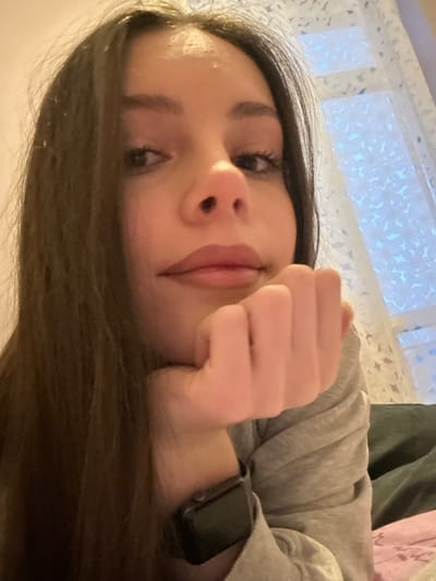 A close-up, slightly overhead photograph depicts a female individual in an indoor setting within Волховец, Russia. The individual has long, dark brown hair and fair skin. Her eyes are open and directed forward, towards the camera, and her lips are visible with a neutral or light-colored product applied. She is wearing a light grey long-sleeved top. Her left hand is fisted, resting beneath her chin or jawline. A dark-colored smartwatch with a square face is present on her left wrist. The background features a plain light-colored wall on the left and a window on the right, covered by a sheer white curtain exhibiting a floral or geometric pattern. Partial views of dark and light pink fabrics are present in the lower right background. Illumination is soft and consistent throughout the scene.