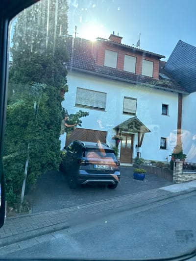 A person is trimming a tree next to a white house in Oberwetz, Germany. The house has a dark-tiled roof and a wooden front door with a small porch. A dark-colored SUV is parked in the driveway. The scene is viewed through a window, which shows reflections and some blur. The sun is shining brightly, casting lens flares.