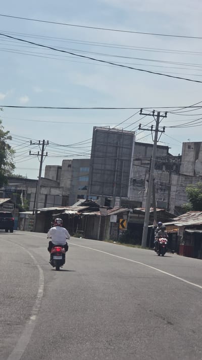 The image depicts a street scene in Lada, Indonesia. A road runs horizontally across the frame. Two motorcycles are present: one in the foreground being ridden away from the viewer and another in the distance on the road. Buildings, including a large billboard structure, line the side of the road. Utility poles and power lines extend across the sky. The weather is sunny and clear.