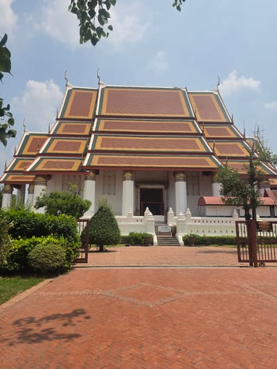 A building with a tiled multi-tiered roof stands in Phra Nakhon Si Ayutthaya City Municipality, Thailand. The roof tiles are rectangular and maroon, trimmed with yellow and green. The building has a white facade with columns. A red brick walkway leads alongside the building. Green shrubbery grows in front of the building. A tree is visible on top of the roof. The sky is light blue with scattered clouds.