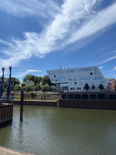 The image shows a modern, angular white building with numerous windows, situated on the bank of a river or canal. The building's architecture is distinctive, with a sloping facade and geometric window arrangements. In the foreground, the murky green water of the waterway is visible, with a dark metal embankment along the edge. To the left of the modern building, there are older buildings and lush green trees. Two tall blue industrial cranes are also present on the left side of the image, suggesting a dock or port area. The sky is a bright blue with scattered white, wispy clouds. The scene appears to be a cityscape, likely in an industrial or waterfront district. This specific building is the "Rheinpark-Center" in Neuss, Germany, situated along the Rhine river. There are no people visible in the image.