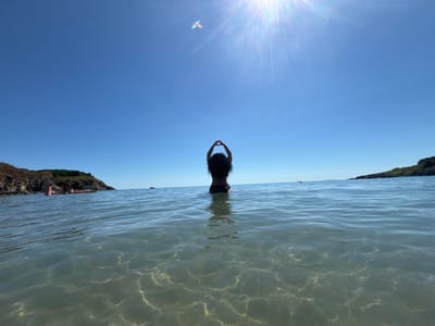 A low-angle, wide-shot photograph captures a person from behind, standing in shallow, clear ocean water with a sandy bottom visible. The person, with long dark hair and wearing dark swimwear, is facing away from the camera and has their arms raised to form a heart shape above their head. The sun is shining brightly in the upper right portion of the frame, creating lens flare and casting shimmering patterns on the water's surface. In the distance, the sea stretches to the horizon under a clear blue sky. On the left side of the image, a rocky, vegetated coastline is visible, with two people in kayaks near the shore. On the right side, another rocky coastline is visible in the distance. The overall scene depicts a serene and sunny day at the beach in Rezovo, Bulgaria.