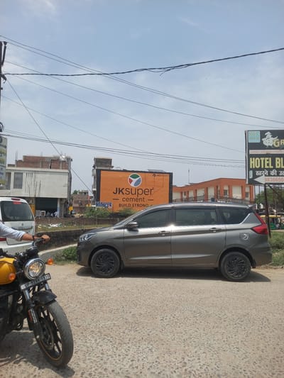 The image shows a street scene in Orai, India. A gray car is parked on the right side of the frame on a paved road. A yellow motorcycle is parked near the car with its front wheel and headlight prominently visible. The motorcycle's license plate reads "UP-92AH 1939". Behind the car, there are several buildings, including one with a large orange advertisement for "JKsuper cement BUILD STRONG". A hotel sign is also visible that says "HOTEL BL", with the text "ठहरने की उत्" and the phone number "9838650". Power lines are strung across the sky. The sky is blue with some clouds.