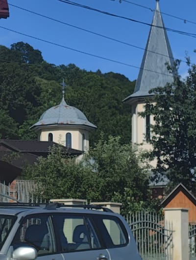 In Vișeu de Jos, Romania, on a clear day, two church towers are visible behind a grey SUV and a metal fence. The tower on the left is round with a light blue dome and a cross on top. The tower on the right is taller and pointed, also with a light blue roof and a cross at its apex. Dense green trees and foliage surround the church structures. The sky is a bright blue with visible power lines crisscrossing the frame. The foreground is occupied by the top portion of a grey SUV and a decorative metal fence.