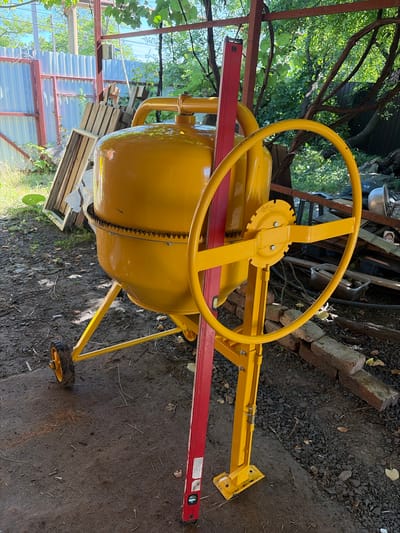 An outdoor, full-shot, medium-angle, eye-level, natural light, documentary-style image shows a bright yellow, portable cement mixer. The mixer is situated on a dirt and gravel surface, with a small portion of a concrete pad visible in the foreground. To the left, a corrugated metal fence is visible, behind which are stacked wooden pallets. In the background, lush green foliage from trees and vines forms a dense canopy overhead, with some sunlight filtering through. A red spirit level is propped against the mixer's rotating drum, indicating it may be in use or undergoing setup. The scene appears to be in a backyard or construction area, possibly on the outskirts of Krugloye, Russia.