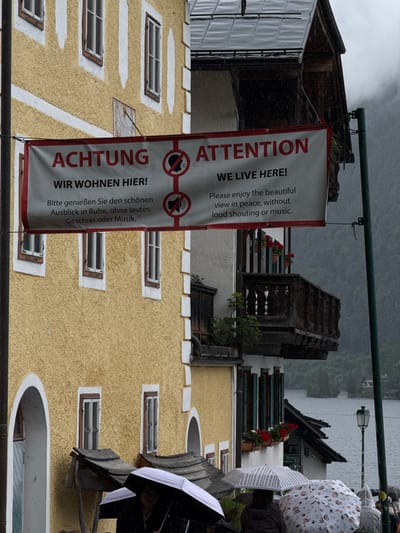 This image captures a street scene in Hallstatt, Austria, on a rainy day. A prominent banner hangs across a building, displaying the text "ACHTUNG WIR WOHNEN HIER!" and "ATTENTION WE LIVE HERE!" in both German and English. The banner also features graphical symbols indicating no loud noises or music. Below the banner, a traditional yellow building with white trim and several windows is visible. In the foreground, people are holding umbrellas, suggesting the ongoing rain. A balcony with red flowers adorns the building to the right of the yellow one. In the background, a body of water, likely a lake, and lush green hills can be seen, characteristic of the Hallstatt landscape. The overall atmosphere is damp and overcast, typical of the moody weather often experienced in this scenic Austrian town.