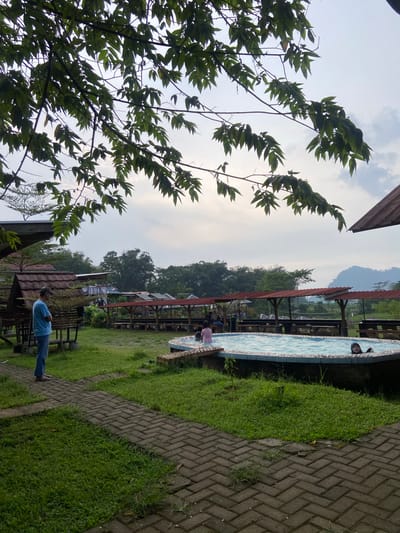 The image depicts a view in Penanggungan, Indonesia, showing a lush outdoor setting. A large leafy tree occupies the left foreground, partially obscuring the scene. In the middle ground, there's a swimming pool with someone in the water, surrounded by grassy areas and brick-paved walkways. Several small structures with red roofs and open sides are visible, suggesting a recreational area. A man in a blue shirt and jeans stands near one of these structures in the lower right of the image. In the background, there's a line of trees and a cloudy sky.