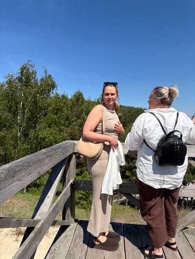Two women are standing on a wooden observation deck. The woman on the left is wearing a tan sleeveless dress and carrying a tan shoulder bag. She has sunglasses on top of her head and is looking towards the camera. The woman on the right is wearing a white oversized shirt and brown pants. She has a black backpack on and is looking away from the camera, talking to the other woman. The deck is made of weathered wood and overlooks a natural landscape with trees and sand dunes. The sky is clear and blue. The location is Morskoye, Russia.