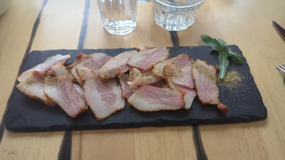 A still life image captures a serving of sliced cured meat on a black rectangular slate platter, situated on a light brown wooden table in Slokoshtitsa, Bulgaria. The platter holds approximately fifteen to twenty thin, irregular slices of meat, each displaying a marbled pattern of pink muscle and white fat. A yellowish-green granular spice is unevenly sprinkled over the meat and the platter's surface. Several dark green leafy sprigs, consistent with arugula, garnish the right side of the platter. In the background, a clear, vertically fluted glass tumbler is positioned to the upper left, and a smaller, clear glass dish with a textured base is to the upper right. A silver knife and a silver fork are partially visible on the table to the far right, suggesting a dining setting. The wooden table exhibits visible grain and darker seams between planks. No individuals are present in the scene.