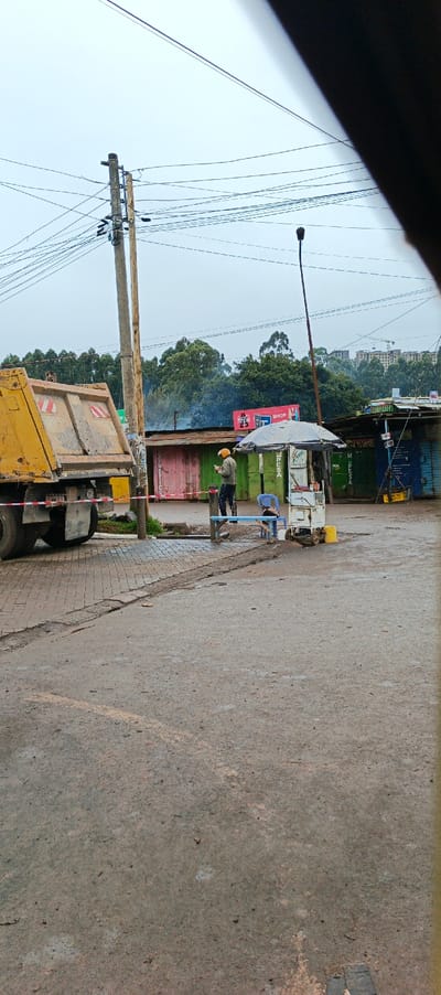 A yellow dump truck sits on the left side of a cracked pavement area. A man in a yellow helmet stands near small, colorful storefronts, possibly using a phone. An umbrella-covered stand with a blue chair is beside him. Utility poles with wires are visible above. In the background, green trees and buildings are seen under a cloudy sky. Part of a vehicle obscures the right edge of the frame.