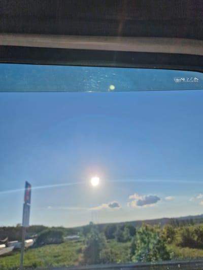 The image captures a bright, sunny daytime scene viewed from inside a vehicle, looking out through a window. The perspective is rotated 90 degrees clockwise from standard orientation. A vast, clear blue sky dominates the view, with the sun positioned centrally, creating a prominent lens flare. A few small, scattered white clouds are visible in the distance.
On the left, the dark interior frame of the vehicle's window is visible, with the blue-tinted glass showing numerous scratches and a small, greenish-yellow circular reflection or flare. Faint, white text, likely manufacturing details or a vehicle identification number, is etched into the upper left corner of the window glass; it is mostly illegible, but fragments like "TOP AS M" and other characters are discernible.
The lower portion of the scene depicts a landscape with a hazy horizon line, suggesting distance. Green trees and bushes line what appears to be the edge of a road or embankment on the right side. A partially visible road sign, with dark lettering on a lighter background, stands upright in the lower right quadrant. The overall impression is of a clear and pleasant day, likely while traveling in Tatarka, Russia. No people are visible in the image.