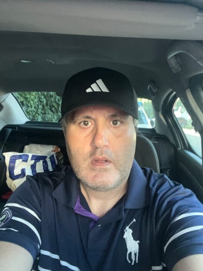 A man is sitting in the driver's seat of a car. He is wearing a black baseball cap with the Adidas logo and a navy blue polo shirt with white stripes on the sleeves and a white Polo Ralph Lauren logo on the chest. The car's interior is visible, including the dashboard, headliner, and rear seats. Behind the man, there are bags and other items in the back seat. The background outside the car shows greenery and a glimpse of another car. The man is looking directly at the camera. The scene appears to be a selfie taken inside a vehicle, likely in a residential or suburban area, given the foliage. There is no specific information within the image that indicates the location is the city of Chantepie, France.