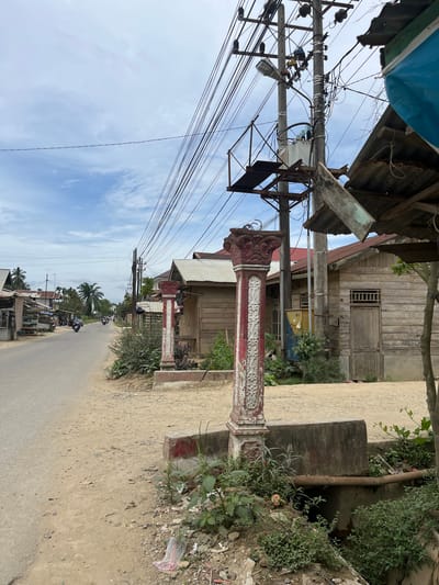 A roadside scene in Durian, Indonesia, depicts a paved road running through a village. To the left, a row of small shops with open fronts displays goods, and several motorcycles are parked. Further down the road, motorcycles with riders are visible. On the right side of the road, a wooden house with a corrugated metal roof stands. A decorative red and white pillar is positioned in the foreground, near a concrete structure and an open drain with a metal pipe. Overhead, a dense network of electrical wires and a utility pole with a transformer dominate the sky. Vegetation, including trees and bushes, is present along the roadside and near the structures. The sky is cloudy with patches of blue.