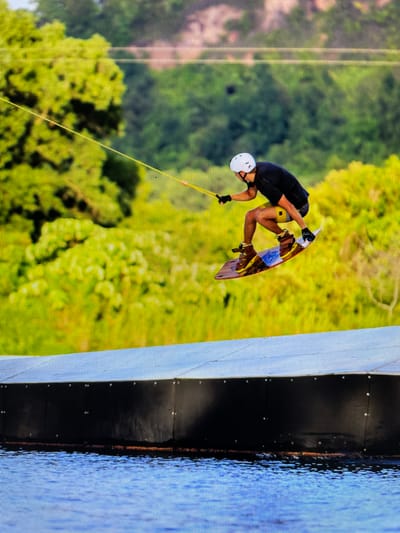 A man on a wakeboard is performing a trick in the air in Bacong, Philippines. He is wearing a white helmet, black shirt, and gloves. He is connected to a rope and is near a dock in the water with green trees in the background.