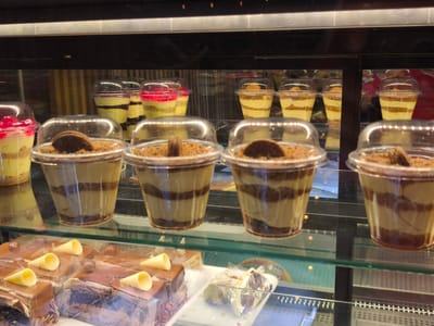 The image displays a close-up view of a dessert showcase, likely inside a bakery or cafe. The scene is indoors, well-lit, and highlights a variety of sweet treats arranged on multiple glass shelves.
In the prominent middle and foreground, several individual parfaits are presented in clear plastic cups with dome-shaped lids. These parfaits feature distinct alternating layers of light-colored cream or pudding and dark brown crumble or chocolate, creating an attractive striped pattern. Many of these are topped with a small, round, dark brown cookie.
On the upper shelves, other types of parfaits are visible. Some have layers of yellow and light green, while others feature a bright red jelly or fruit topping over yellow layers, also contained in clear plastic cups.
The bottom shelf showcases multiple slices of rich chocolate cake or pastry. These rectangular pieces are covered with a smooth, glossy chocolate glaze and are decorated with small, light yellow cone-shaped elements and tiny chocolate spheres. Some of these cake slices appear to be individually wrapped in clear plastic.
The display case itself is dark-colored, with clear glass shelves and partitions, exhibiting reflections from the indoor lighting. The arrangement of the desserts is neat and organized, intended for sale. There are no people visible in the image, nor is there any discernible text. The time of day and weather cannot be determined from the indoor setting. The image provides no specific visual cues to its location, though it is contextualized as Dagonovo, Bulgaria.