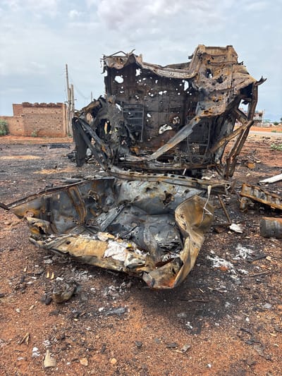 The image depicts a scene of destruction in Um Laba, Sudan. In the foreground and mid-ground are the heavily charred and mangled remains of what appears to be a vehicle, likely an SUV or van, completely destroyed by fire. The metal is twisted, melted, and blackened, with numerous holes and gaps visible, suggesting the intensity of the blaze and possible impacts. Some white, fibrous material, possibly insulation or debris, is scattered around the wreckage. The ground is covered in reddish-brown soil, ash, and scattered debris from the destruction.
In the background, a damaged brick building with visible holes, likely from gunfire or shrapnel, stands to the left. A utility pole with electrical wires is also visible. Further in the distance, other damaged structures and the landscape of Um Laba can be seen under a cloudy sky. The overall impression is one of recent conflict or disaster, with the destroyed vehicle being the central subject of the image. No people are visible in the frame.