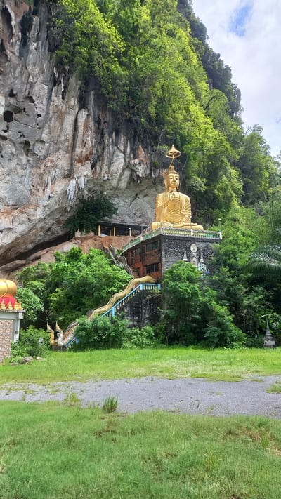 The image shows a golden Buddha statue at Wat Tham Ta Pan temple in Thap Put, Thailand. The Buddha is located on the side of a limestone cliff covered with lush green vegetation. A staircase with a dragon statue railing leads to the statue. A building is behind the statue. A grassy area and pathway are in the foreground. The sky is visible above.