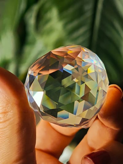 A close-up, eye-level, indoor shot shows a person holding a faceted crystal ball with their thumb and forefinger. The crystal ball is clear with rainbow-like refractions, suggesting it is interacting with light. The background is a blurred image of green foliage, possibly a houseplant. The person's skin tone is fair, and their fingernails are painted a dark red. The scene does not contain any identifiable landmarks or elements that would indicate a specific location in Kursk, Russia, or any city. The lighting is bright and focused on the crystal ball.