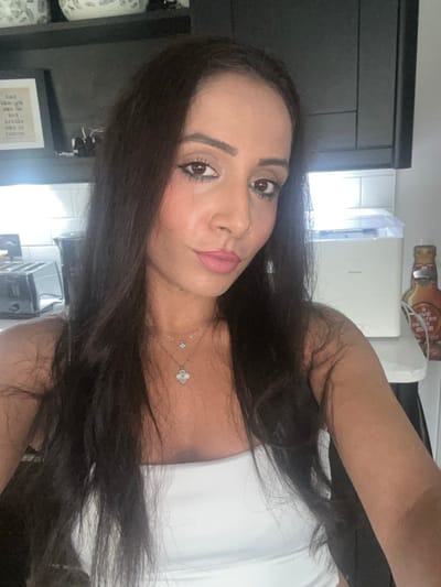 The image depicts a woman with long, dark hair and light brown skin, presenting a direct gaze into the camera with a slight smile. She exhibits dark eyes with visible eyeliner and mascara, pink lipstick, and a small nose ring on her left nostril. She is wearing a white strapless top and two layered necklaces: one a thin chain with a small blue charm, and another featuring a white four-leaf clover pendant.
The scene is an indoor environment, identified as a kitchen or utility area located in Rushden, United Kingdom. The background features dark overhead cabinets and white subway tile backsplash. On the left side of the frame, a silver two-slice toaster and a dark cylindrical appliance are situated on a light-colored countertop. Above these, a framed print, partially obscured, displays the text "...and though she be but little one is fierce." On the right, a light-colored countertop supports a compact, light-colored appliance, consistent with a countertop ice maker, and a wall-mounted, vintage-style bottle opener with red and yellow graphics and partial text, possibly "HAVE A BAVARIAN BEER." The woman appears to be taking a selfie.