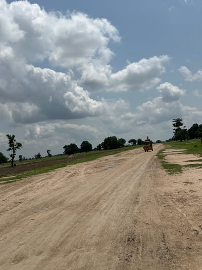 The image depicts a wide, dirt road in Garin Dogo, Nigeria, under a cloudy sky. The road, with visible tire tracks, stretches into the distance, bordered by green fields and scattered trees on the left. A yellow construction vehicle, possibly a backhoe, is seen moving away from the viewer on the road. The sky is filled with dramatic, large white and gray clouds, suggesting either impending rain or recent weather activity. The lighting suggests it is daytime, likely late morning or early afternoon. The overall scene conveys a sense of rural activity and open landscape. There is no visible text in the image.
