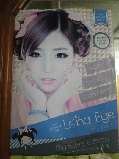 This image depicts a close-up photograph of a consumer product package, located in Keo Kouang, Laos. The primary subject is a rectangular box or display card for "Lena Eye" soft cosmetic contact lenses, specifically the "Big Eyes Edition" in "BROWN COLOUR." The packaging specifies a "6 (SIX) MONTHS SUPPLY" and describes the lenses as "SOFT, THIN, COMFORT."
The front of the package features a portrait of a young East Asian female model. She is looking directly forward, with dark hair styled into two prominent buns or coils, adorned with light pink bows. Her eyes are accentuated with dark eyeliner and appear to be enhanced by dark brown or black cosmetic contact lenses. Her lips are slightly parted, colored with light pink lipstick. Her hands are positioned beneath her chin, showcasing light blue nail polish on her fingernails and a light blue beaded bracelet on her left wrist. She wears a light-colored garment, possibly pink.
The background of the package artwork is a light gray and white geometric pattern. A prominent blue band crosses the lower section of the package, displaying the brand name "Lena Eye" with a stylized crescent moon and stars, and the text "BY: S.H.E." A cartoon character, depicting a girl with pigtails and a bow, is positioned on the lower-left of this blue band. Additional text in Korean, "갈색" (brown color) and "고품질" (high quality), is present.
The photograph itself captures the package partially framed on its left and top-left by a vertical, light brown wooden surface, exhibiting visible wood grain. The right and bottom edges of the image reveal glare and a slightly blurred, lighter background, consistent with an indoor environment. The package exhibits minor surface imperfections or dust.