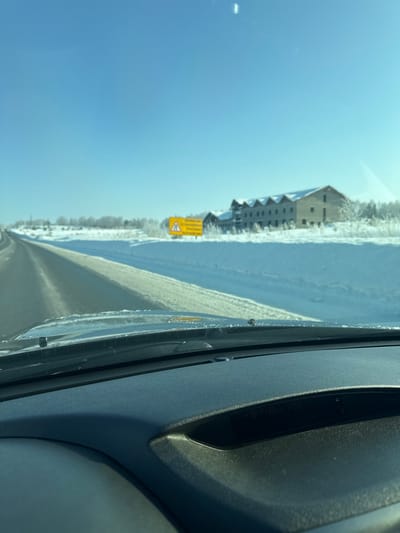 A view from inside a vehicle reveals a snow-covered landscape. A road runs alongside a snow-laden field, bordering a building complex. A yellow warning sign stands prominently along the roadside. A clear, blue sky dominates the upper portion of the frame. The right side showcases the interior of the car, including part of the dashboard and windshield.