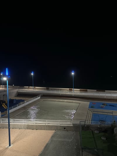 The image shows a night scene of an outdoor swimming pool complex and playground area adjacent to the sea in Saint-Valery-en-Caux, France. The sky is dark, indicating nighttime. Several blue lampposts illuminate the area. The swimming pool is partially filled with water, and the surface reflects the artificial light, creating shimmering patterns. A white railing encloses the pool area. Adjacent to the pool is a blue-surfaced playground with various play structures, including swings and what appears to be a roundabout. Further in the background, a paved walkway runs along the sea wall, lined with wooden benches. The sea is visible as a dark expanse beyond the walkway. There is one person visible, standing near the walkway, and a small white object that could be a bird in flight over the sea. The foreground shows a textured concrete or stone pathway. The overall scene depicts a public recreational area at night.