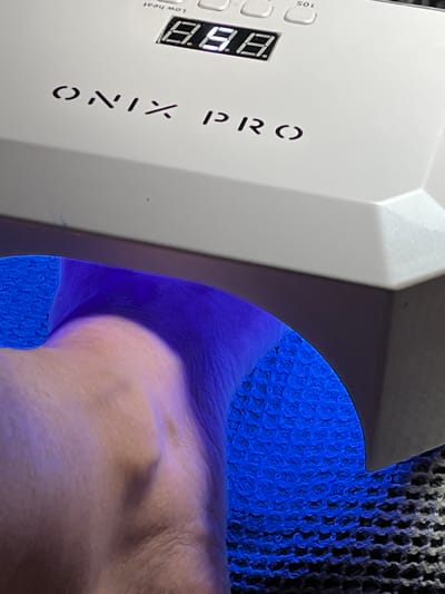 The image shows a close-up of a white UV nail lamp with the brand name "ONIX PRO" printed on it. A digital display on the lamp shows "8.8.". The lamp is emitting a bright blue light, which is illuminating a person's hand and wrist. The hand appears to be resting inside the lamp, likely undergoing a gel nail curing process. The background consists of a textured surface, possibly a mat or a fabric, with a blue woven pattern visible underneath the light. The scene is indoors, and the lighting is dominated by the blue glow of the UV lamp. There is no information available about the specific location in Montrond-les-Bains, France.