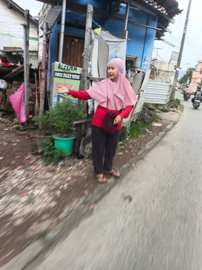 A female individual stands on the unpaved shoulder of a paved road in Dayeuhkolot, Indonesia. She is wearing a pink hijab, a long-sleeved red top, dark trousers, and light-colored sandals. A dark red cross-body bag is slung across her chest. Her right arm is extended forward with an open palm, suggesting a gesture or hailing motion, and her mouth is slightly open. Behind her is a light blue, dilapidated building or shack constructed with wood frames and some corrugated metal. To the left of this building, an older structure with exposed bricks and a pink plastic bag is visible. A green sign with white text, including "AGEENA" and "0813 2022 1010", is affixed to a wooden pole near the blue building. A green potted plant rests on the ground near the woman's feet, amidst dirt and scattered debris. Further down the curving road, two individuals on motorcycles are visible, moving away from the camera. The sky is overcast. The scene depicts a roadside environment with modest residential structures.